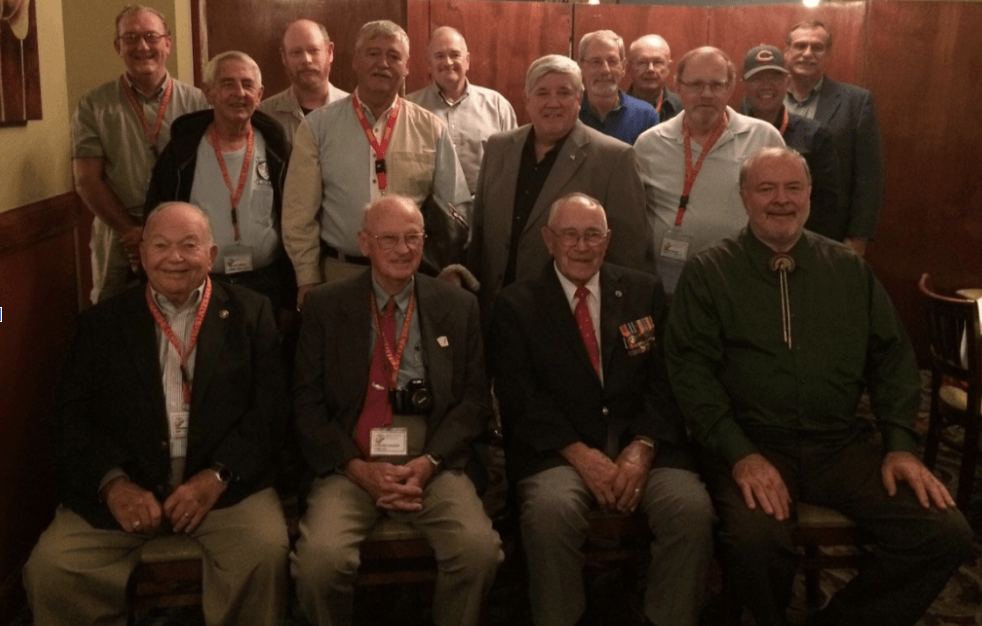 Group of men, mostly older, posing indoors. Some seated in front row, others standing behind them.