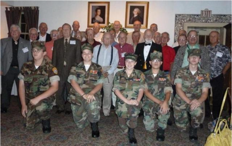 Group photo: Veterans and young cadets posing indoors, portraits in background. Cadets kneel in uniform.