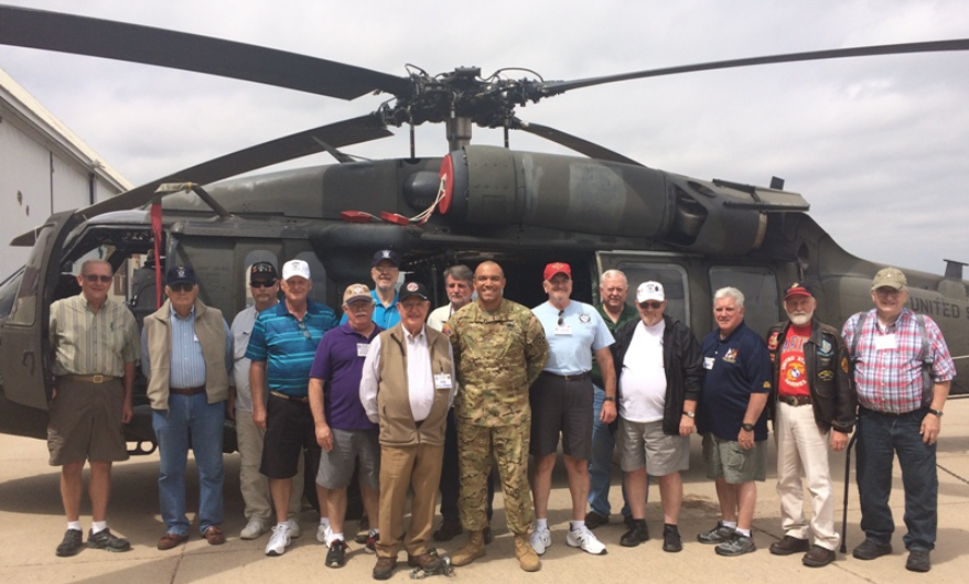 Group of men pose with a Blackhawk helicopter on a tarmac, some wearing veterans' attire.