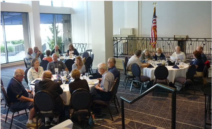People seated at round tables in a brightly lit room with windows, possibly at a luncheon or meeting.