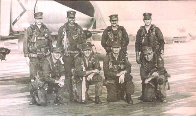 Group of military personnel in flight suits, posing in front of a plane on a runway.
