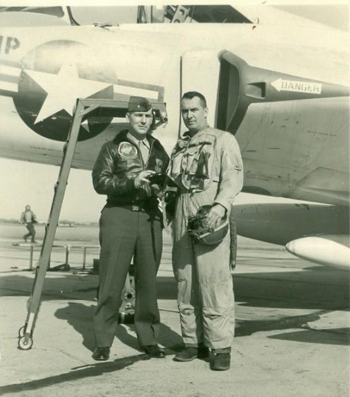 Two men in flight suits stand by a military aircraft. One holds a helmet. Outdoors, sunny.