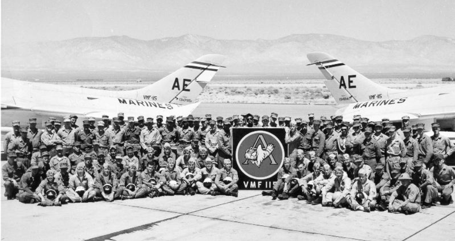 Group of military personnel posing with two aircraft, desert backdrop, unit emblem in foreground.