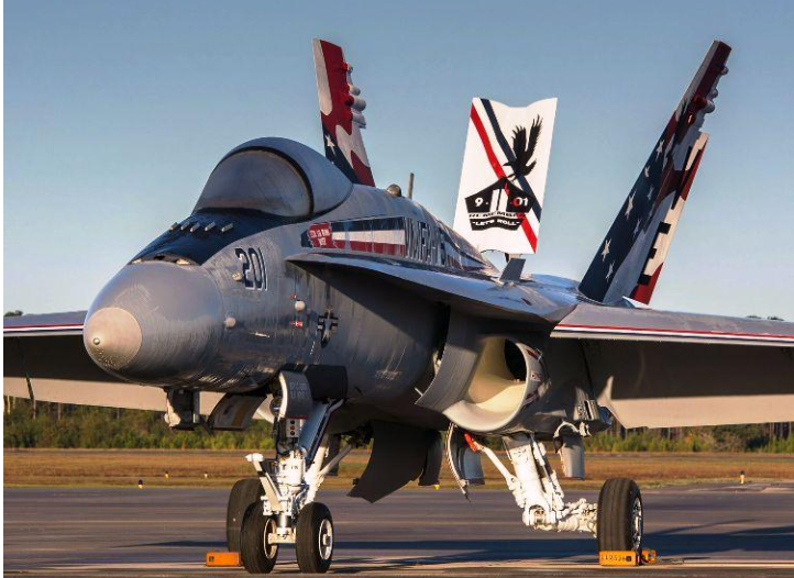 A grey U.S. Navy F/A-18F Super Hornet fighter jet sits on a runway. The tail has red, white, and blue accents.