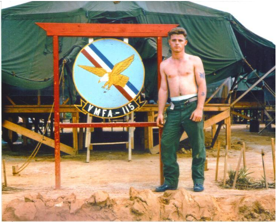 Man standing near a sign with an eagle emblem, in front of a tent; outdoor setting.