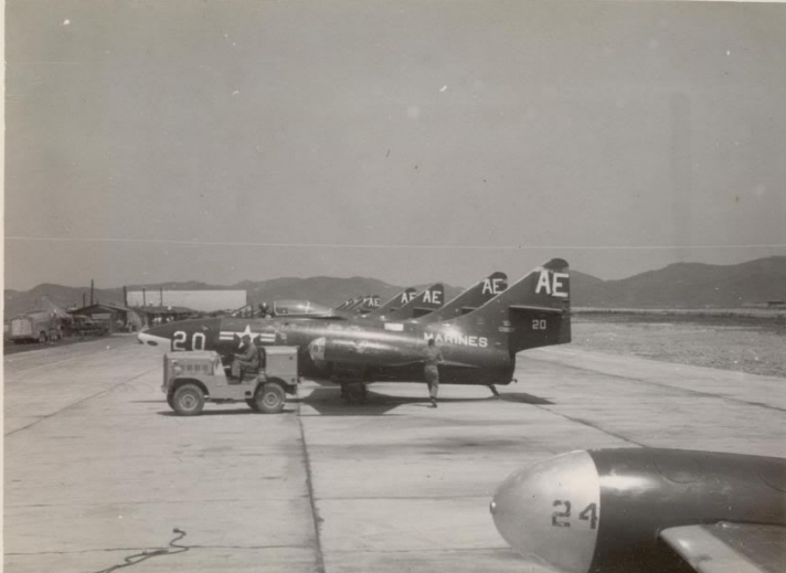 Black jets lined up on a runway, a small jeep parked in front. Mountains and sky in the background.