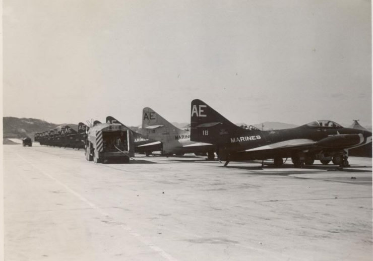 Several dark-colored jet aircraft parked on a runway with a transport vehicle and distant vehicles. 