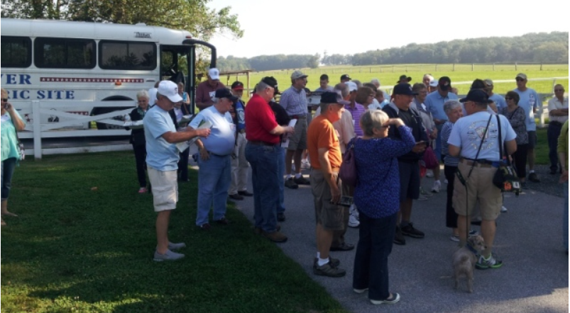Group of people standing near a bus at a historic site, listening to a presentation.