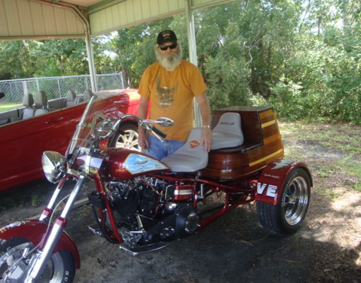 Man with a white beard poses beside a custom maroon motorcycle trike. Outdoor setting, red car visible.