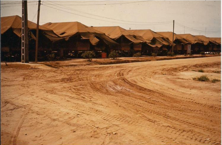 Row of canvas-roofed buildings along a dirt road, likely a military or temporary camp. Sandy, brown ground.