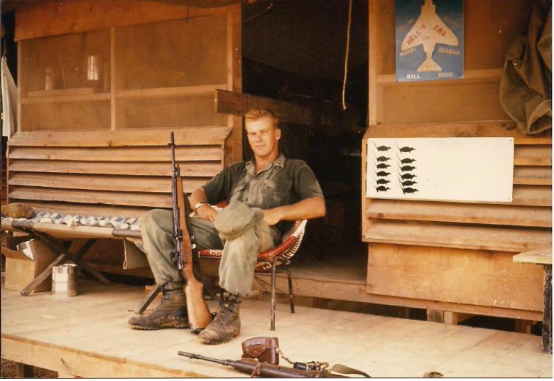 Soldier seated outside a wooden building, holding a rifle, wearing military attire.