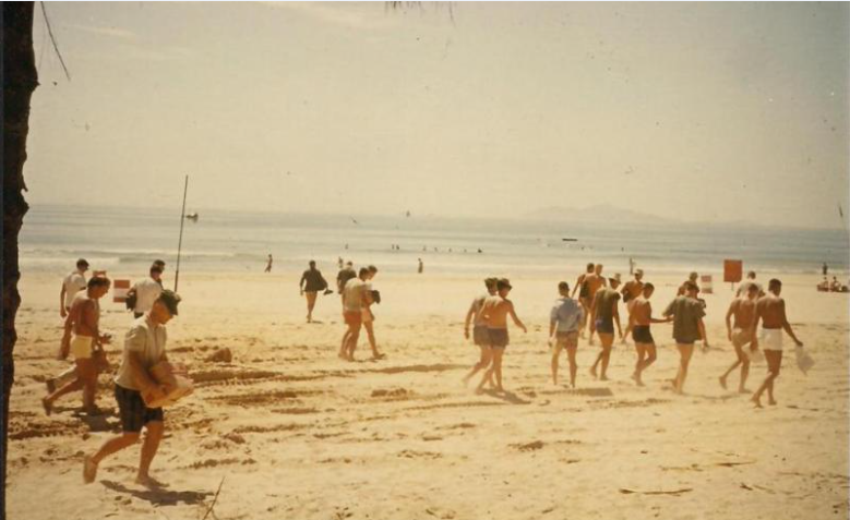 People walking on a beach on a sunny day. The ocean and mountains are in the background.