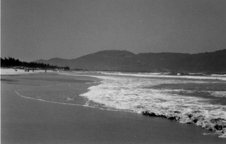 Waves crashing on a sandy beach with mountains in the background under a cloudy sky.