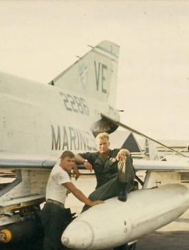Two men pose next to a Marine jet. One sits on a fuel tank, the other leans against the wing.