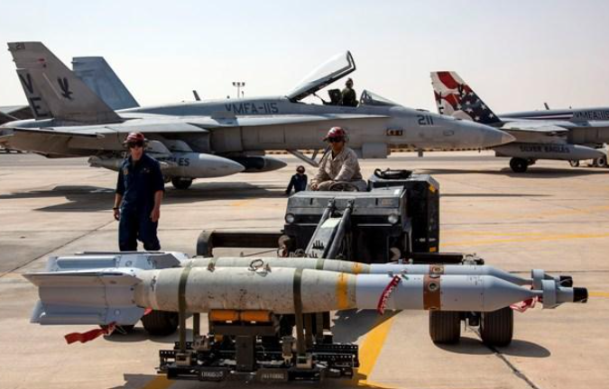 F/A-18 fighter jet on tarmac with technicians, munitions cart carrying a missile.