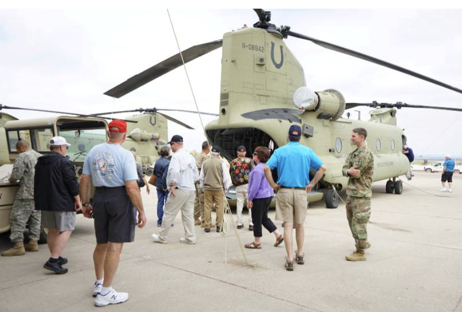 People gather around a military Chinook helicopter on an airfield. Some are looking inside, and a soldier stands nearby.