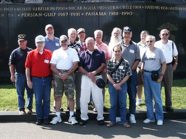Group of men posing in front of a memorial wall inscribed with military conflicts.
