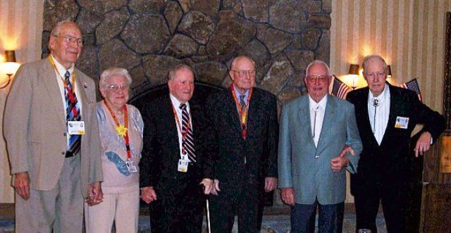 Group of six people posing in front of a stone fireplace. All are wearing jackets and lanyards.
