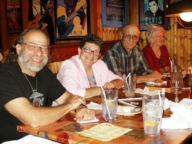 Four people smiling at a table in a restaurant. Posters on the wall, drinks, and food present.
