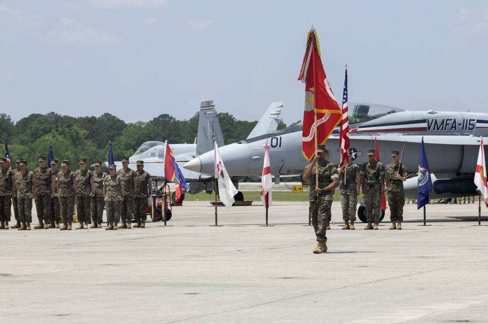 Marines in formation, holding flags, on an airfield. Two fighter jets in background. Sunny, outdoor setting.