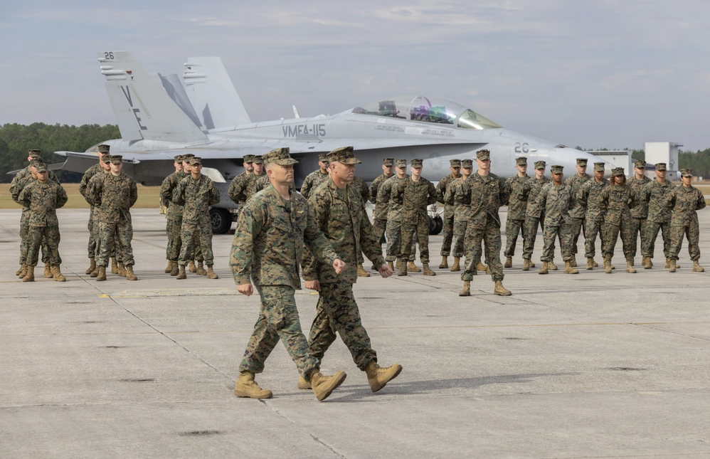 Two uniformed Marines walk; others stand in formation by a fighter jet on a tarmac.