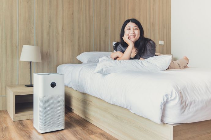 Indoor Air Quality — Memphis, TN — Absolute Comfort Woman lying on a bed with an air purifier, smiling, light-colored bedroom.