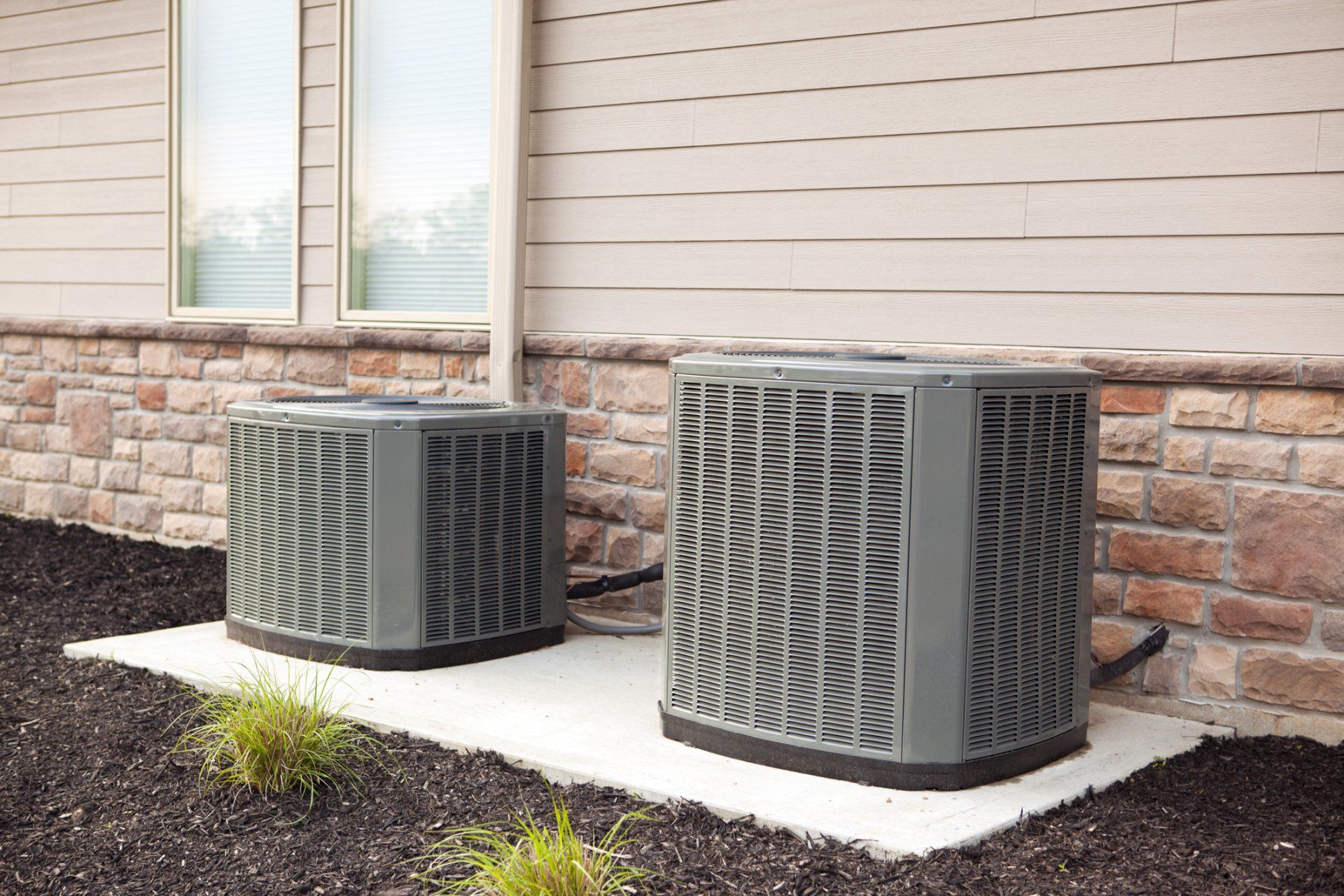 Two Air Conditioning Unit — Memphis, TN — Absolute Comfort Two gray air conditioning units on a concrete pad next to a house with beige siding and brick.