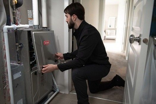 A person kneels in a utility closet, opening the metal service panel of a Bryant HVAC furnace unit. A person kneels in a utility closet, opening the metal service panel of a Bryant HVAC furnace unit.