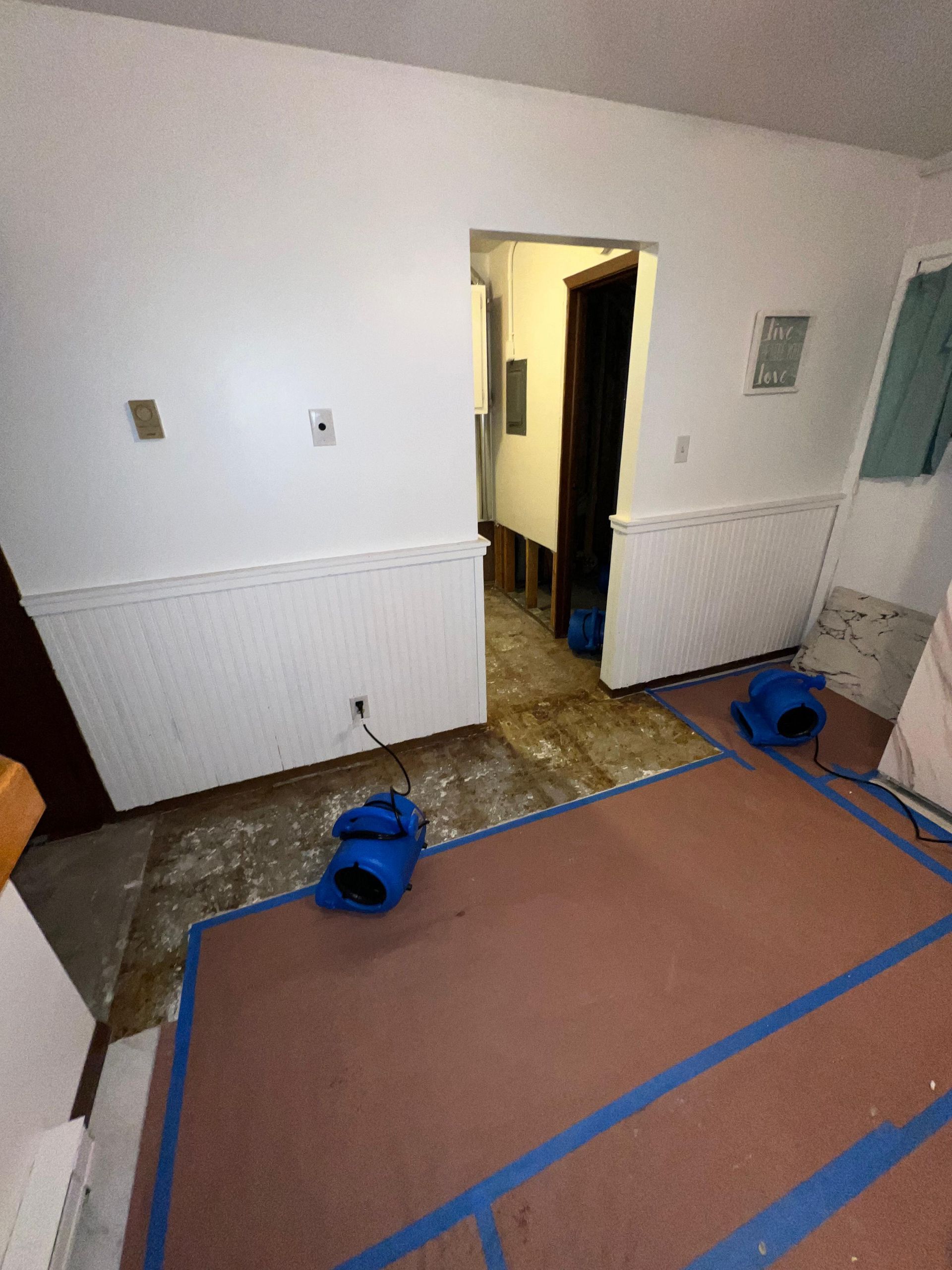 Interior room with white walls, open doorway, taped floor covering, and blue equipment on the floor.