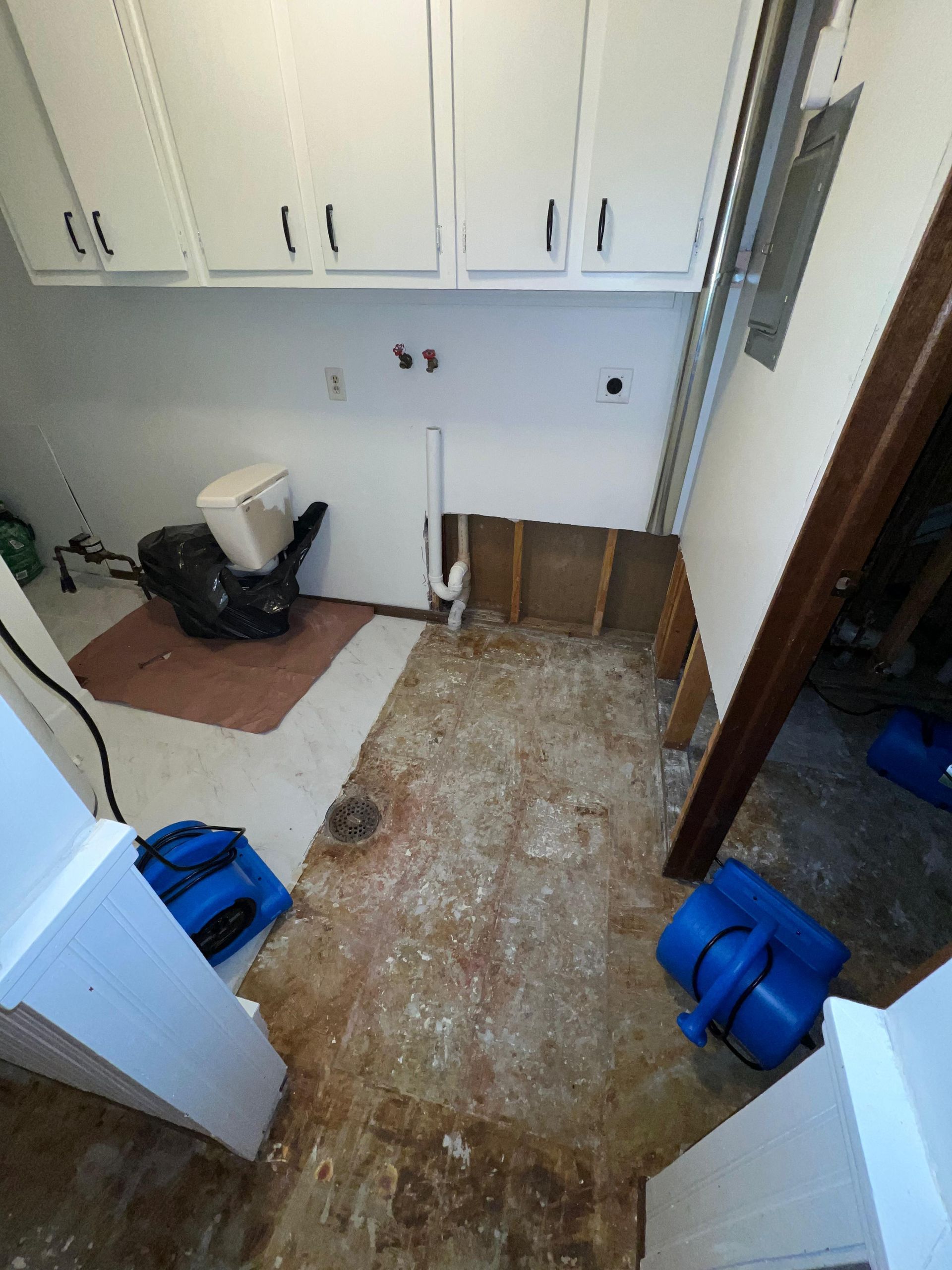 Small laundry room stripped to subfloor, with white cabinets, tools, and blue bags on the floor.