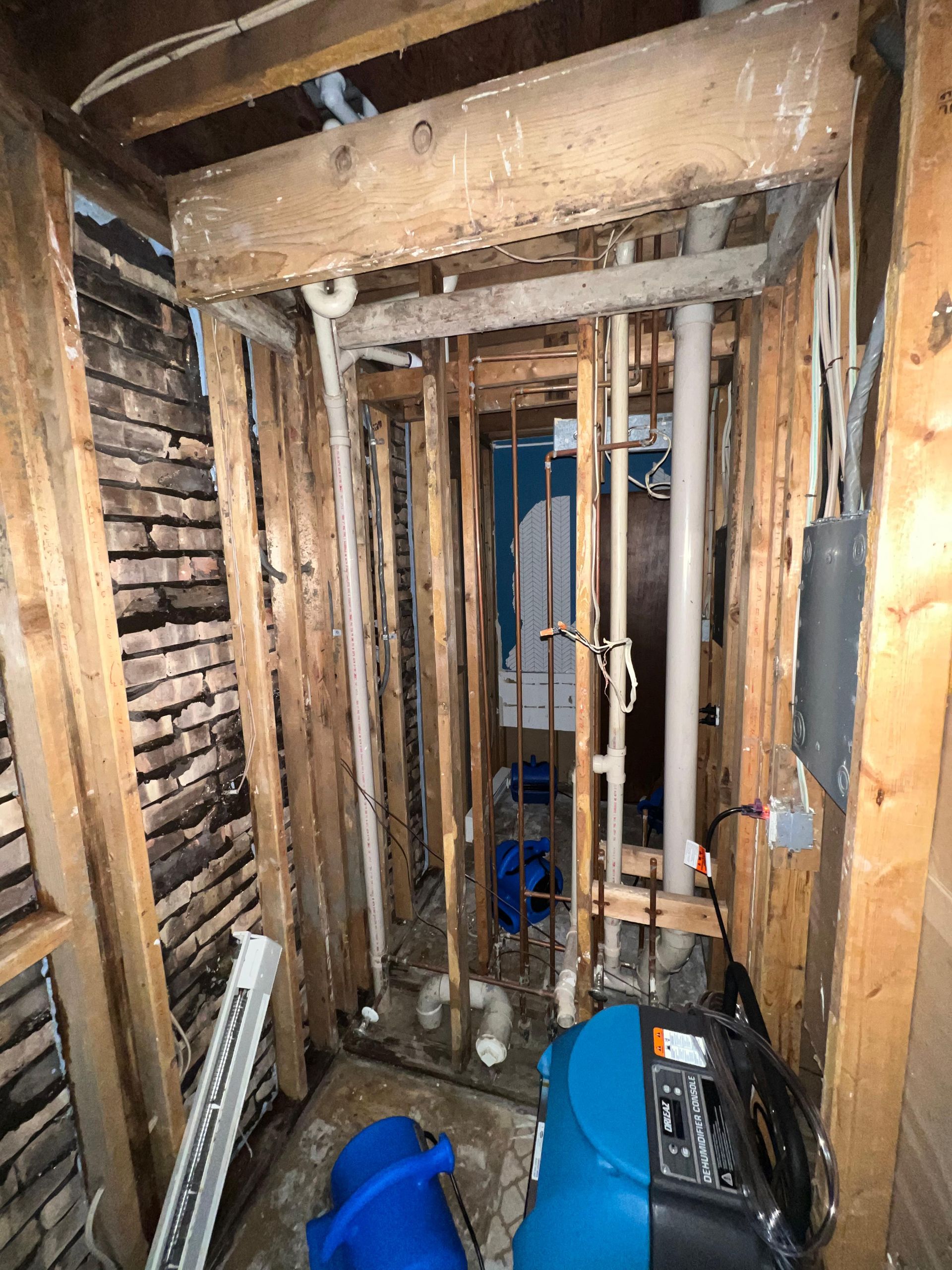 Damaged interior wall framing with exposed studs, pipes, and debris in a narrow renovation space.