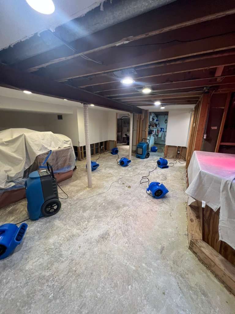 Basement room under renovation with blue fans, covered furniture, and construction debris on the floor