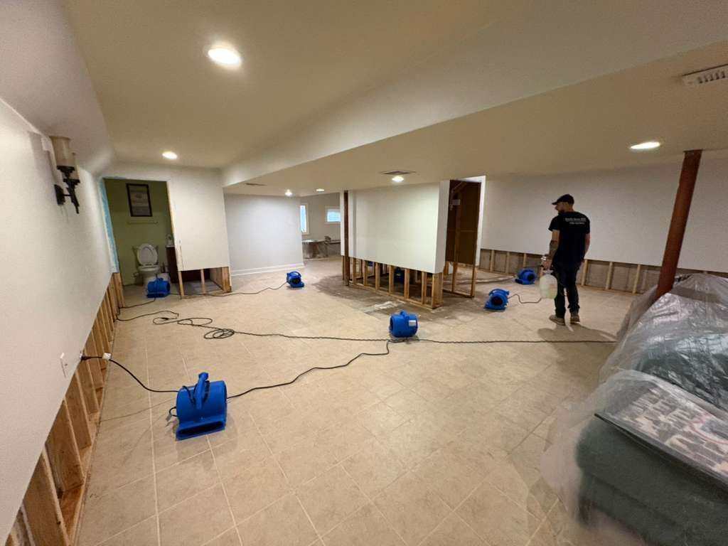 Flooded basement under cleanup, with standing water, hoses, pumps, and a person working near the back.