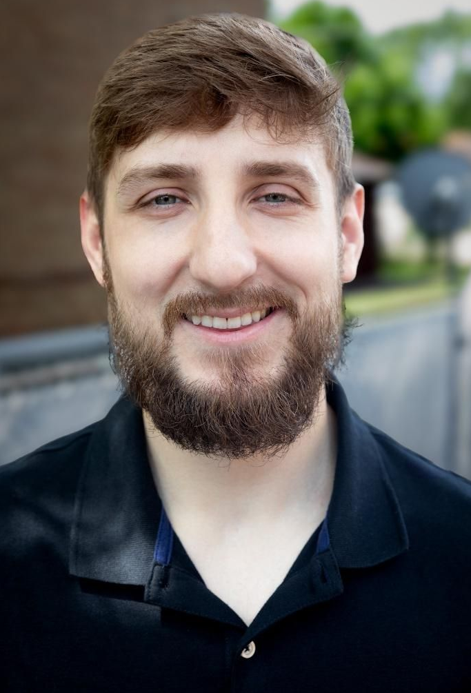 Smiling bearded person in a dark shirt, standing outdoors with blurred greenery in the background
