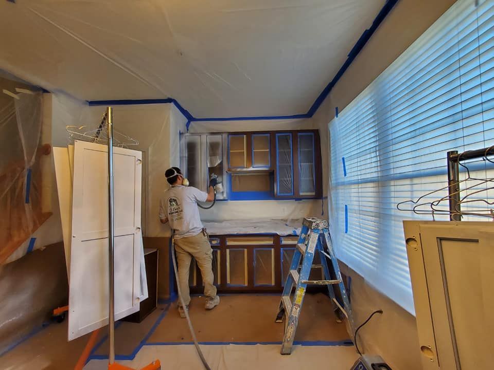 Person painting kitchen cabinets in a room masked with plastic and blue painter’s tape.