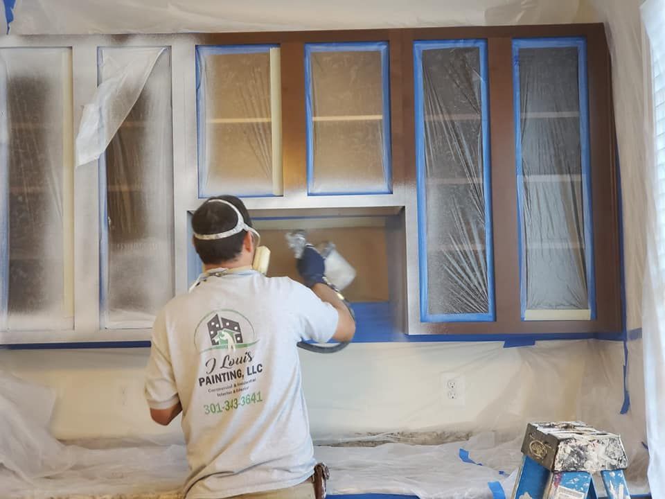 Person spray-painting window trim and wall edges in a room covered with protective plastic and tape.