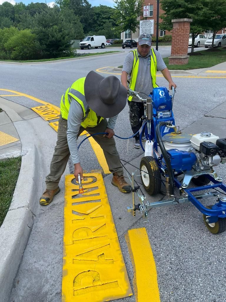 Workers repainting bright yellow curb lettering beside a sidewalk and parking lot with a line striping machine