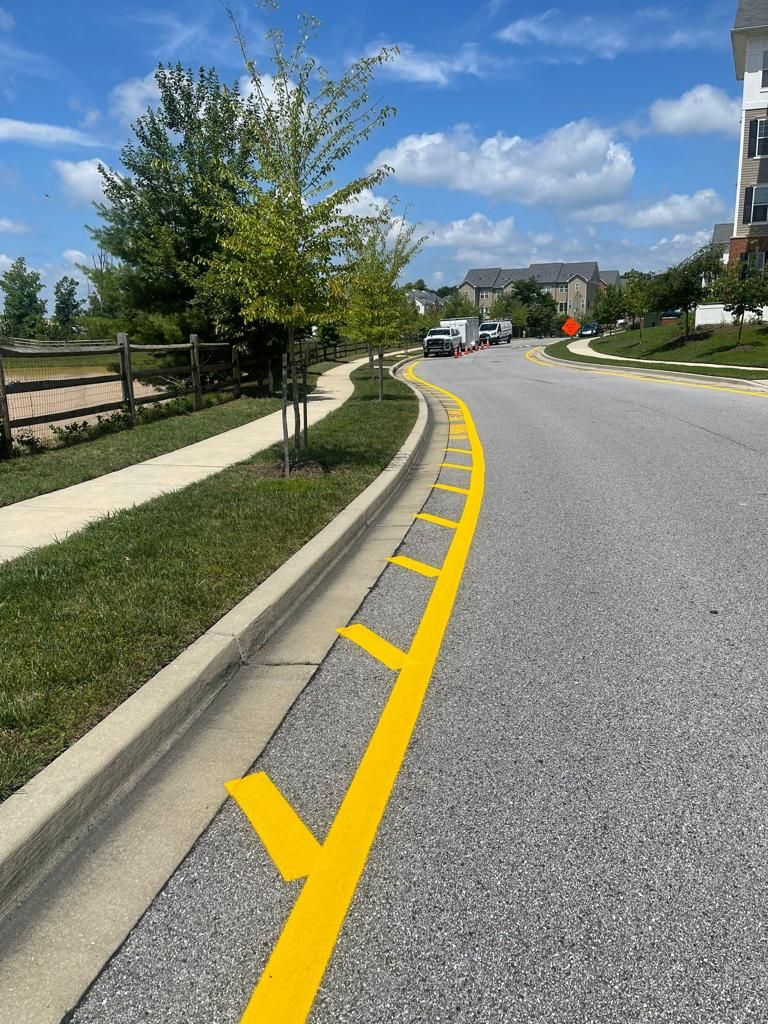 Curved suburban road with yellow curb markings, sidewalk, trees, and houses under a blue sky with clouds