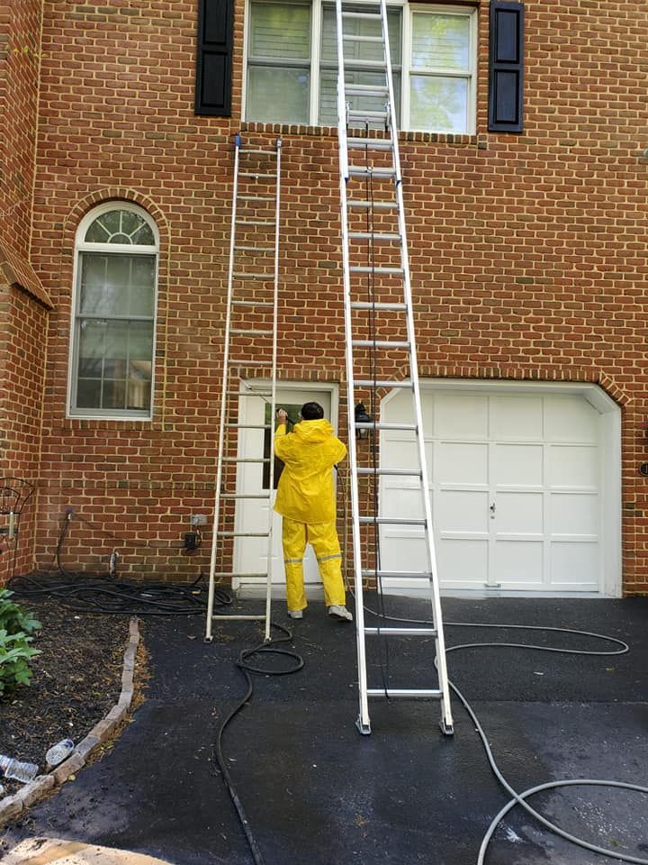 Person in yellow rain gear using a ladder to wash second-story windows on a brick house.