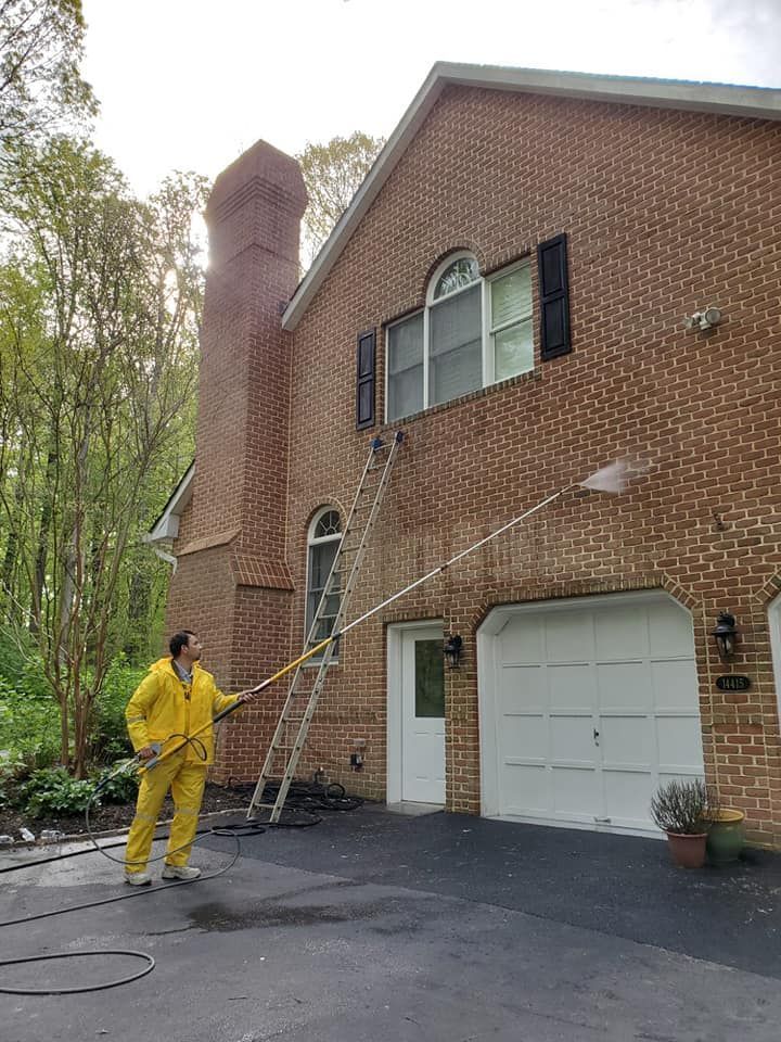 Worker in yellow suit pressure-washes a brick house exterior near a garage and ladder.