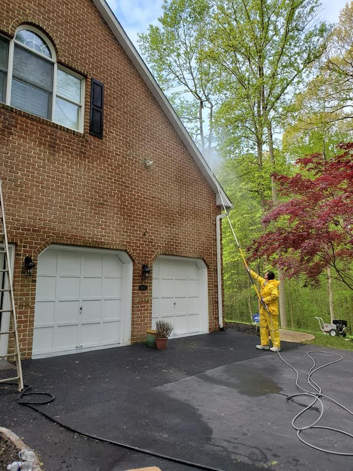 Person in yellow on a driveway beside a brick house, pressure washing near garage doors and trees.