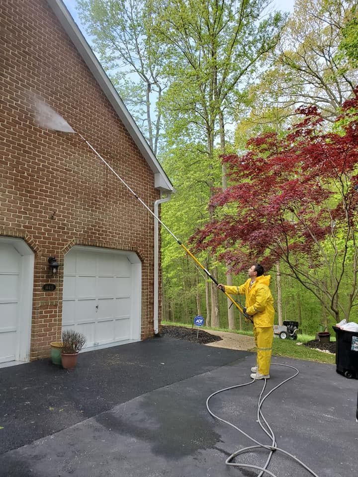 Person in yellow rain gear spraying a red-leaf tree beside a brick house and driveway.