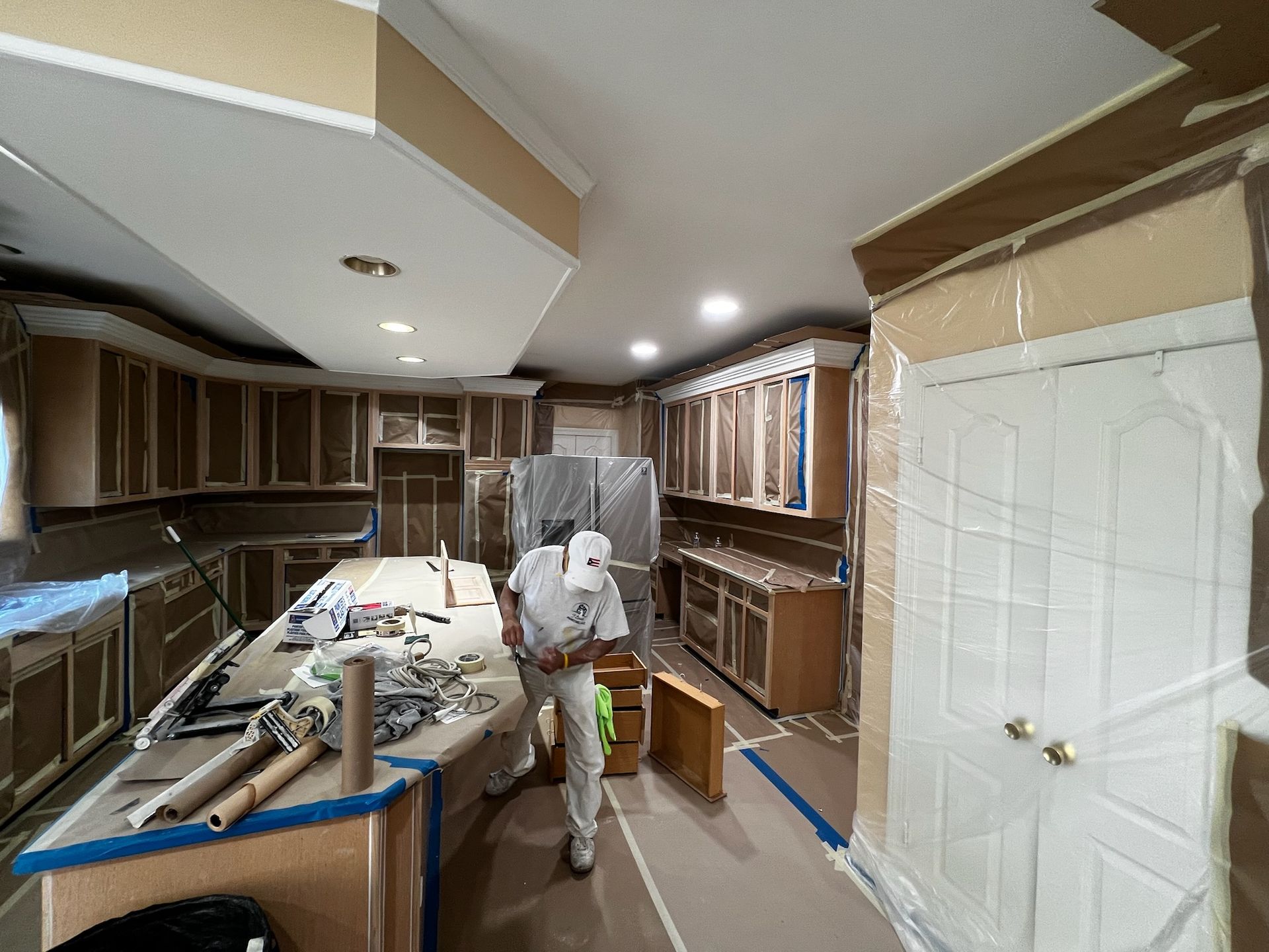 Worker standing in a partially renovated kitchen with cabinets, tools, and plastic-covered surfaces.