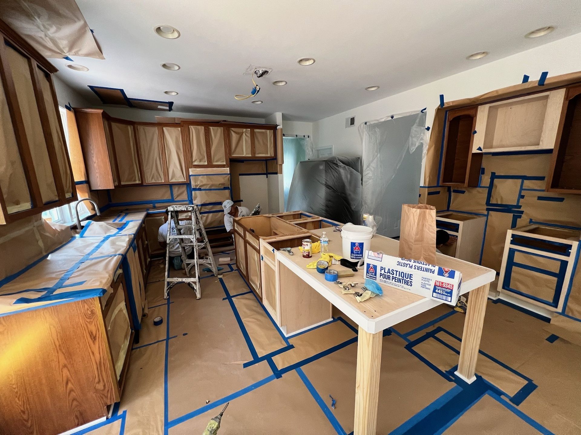 Kitchen under renovation with cabinets taped off, tools on counters, and blue painter’s tape on the floor