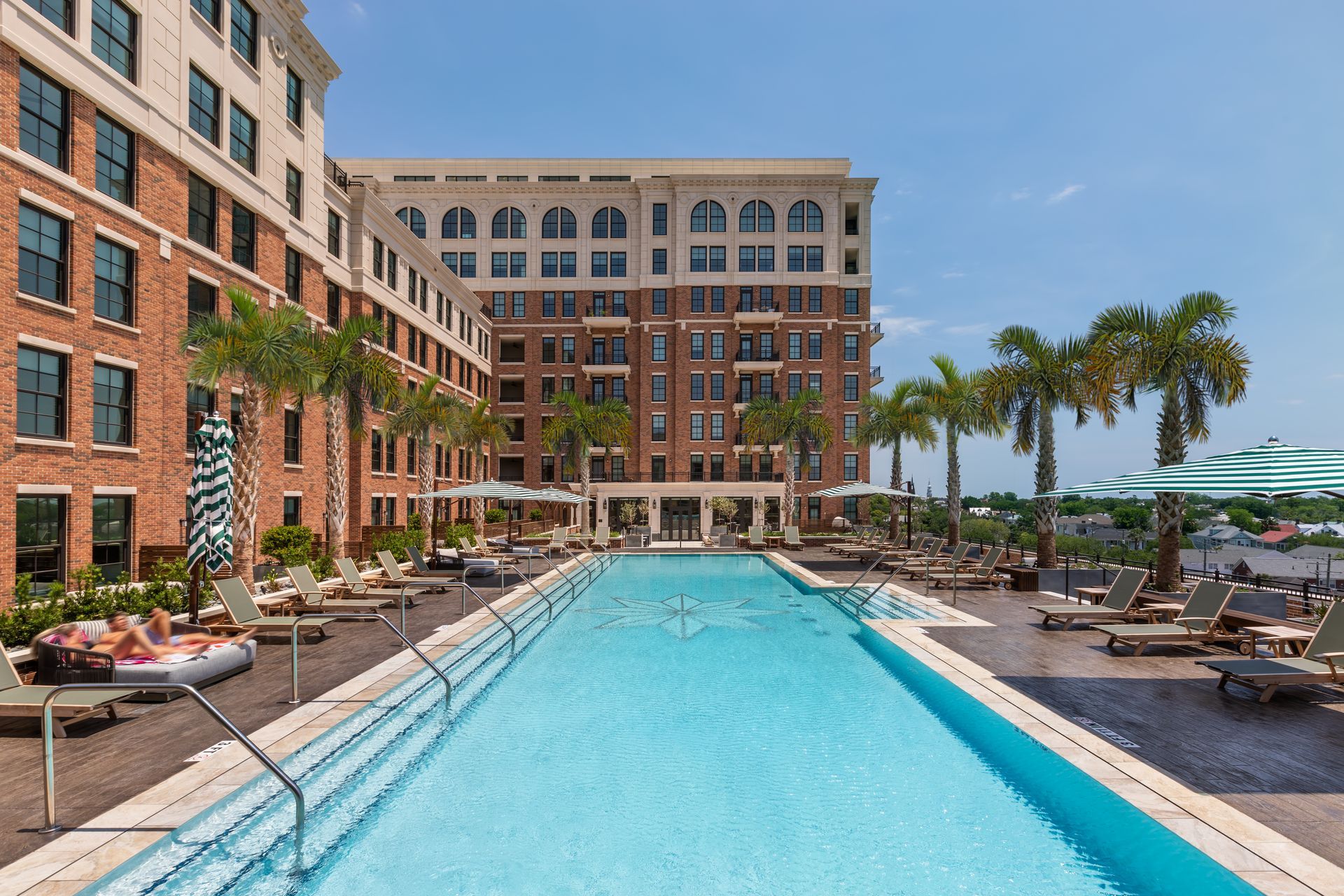 A large swimming pool is surrounded by chairs and umbrellas in front of a large building.
