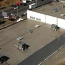 Aerial view of a flat industrial rooftop with HVAC units and a white wall, beside a road with parked trucks.