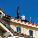 Two workers on a roof repairing red tiles under a clear blue sky.