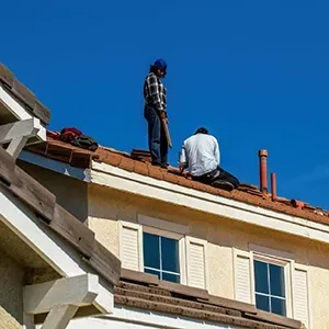 Two workers on a roof repairing red tiles under a clear blue sky.