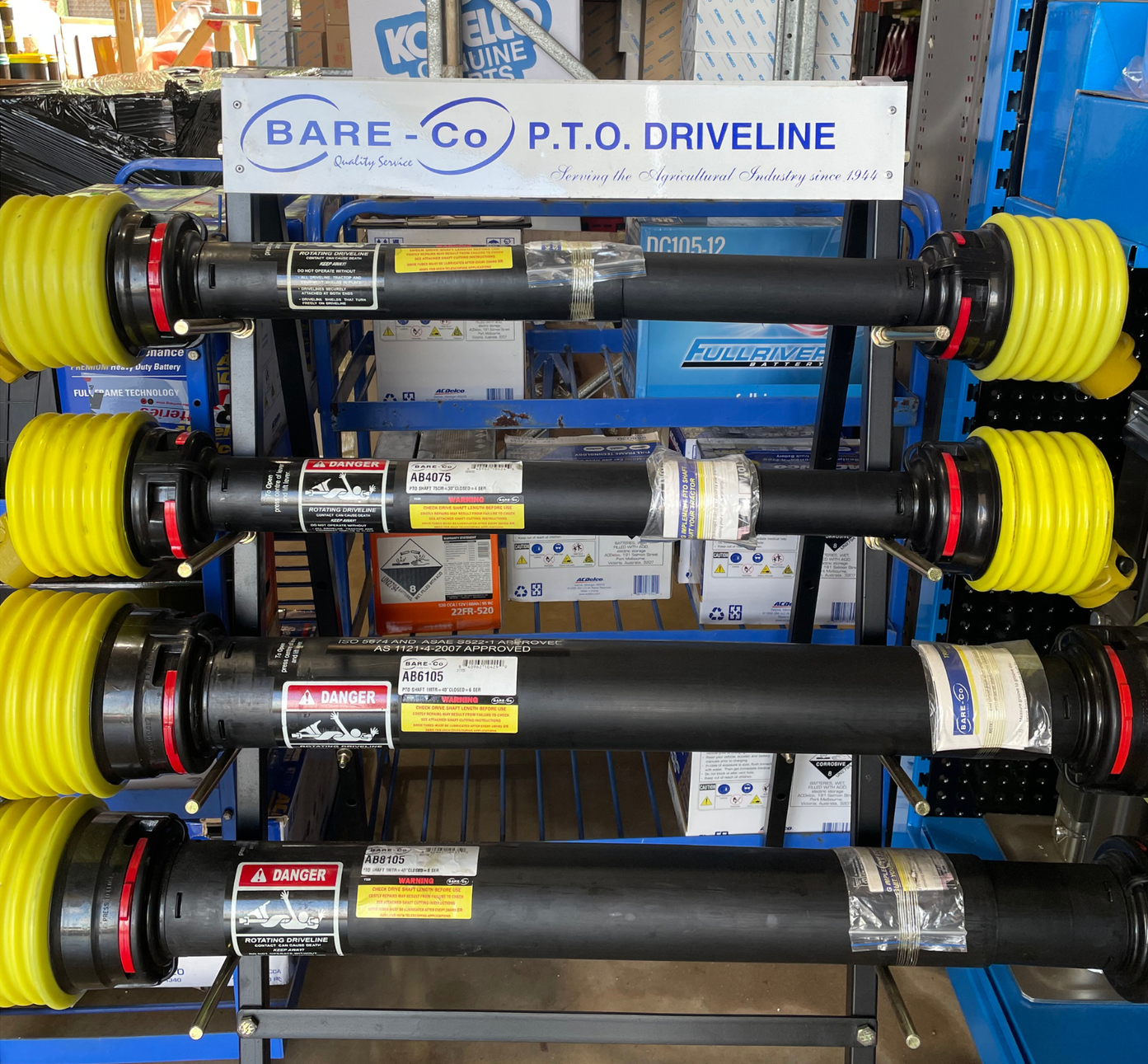 A metal display rack holds four BARE-Co P.T.O. driveline shafts with yellow protective guards, arranged one above the other — Forecast Machinery in Yarrawonga, NT