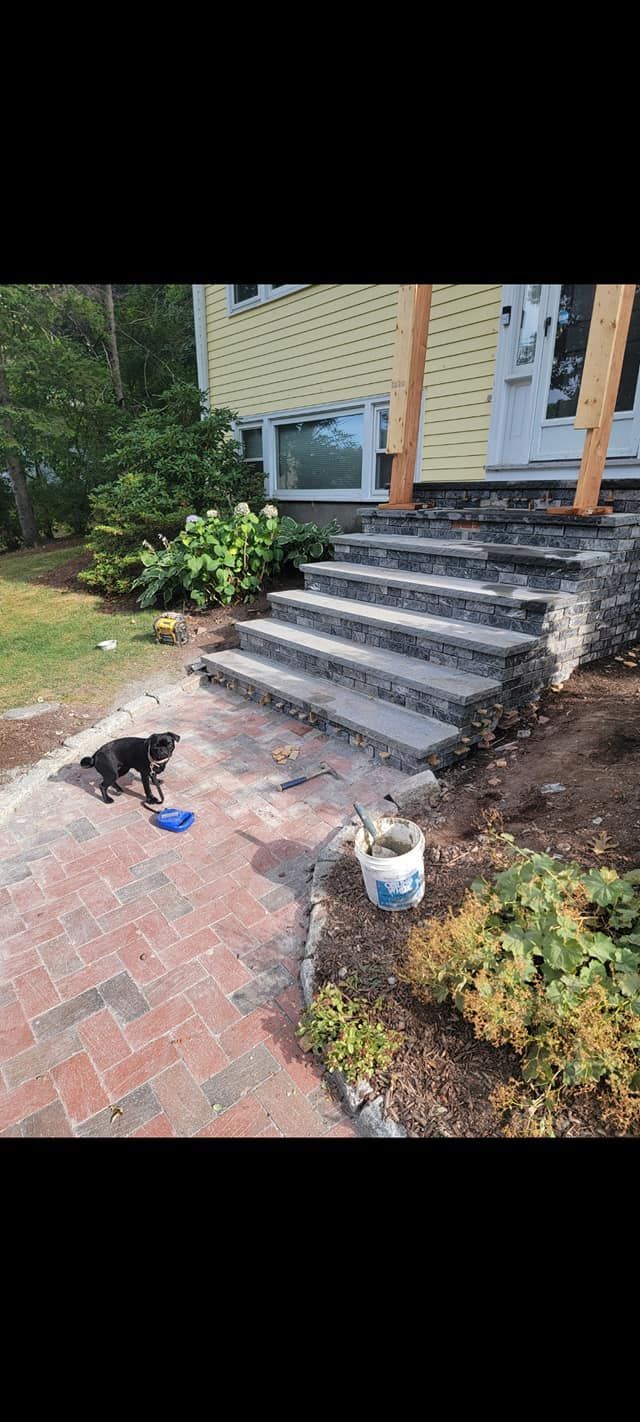 A black dog on a brick patio looks towards stone steps leading to a yellow house with a white door.